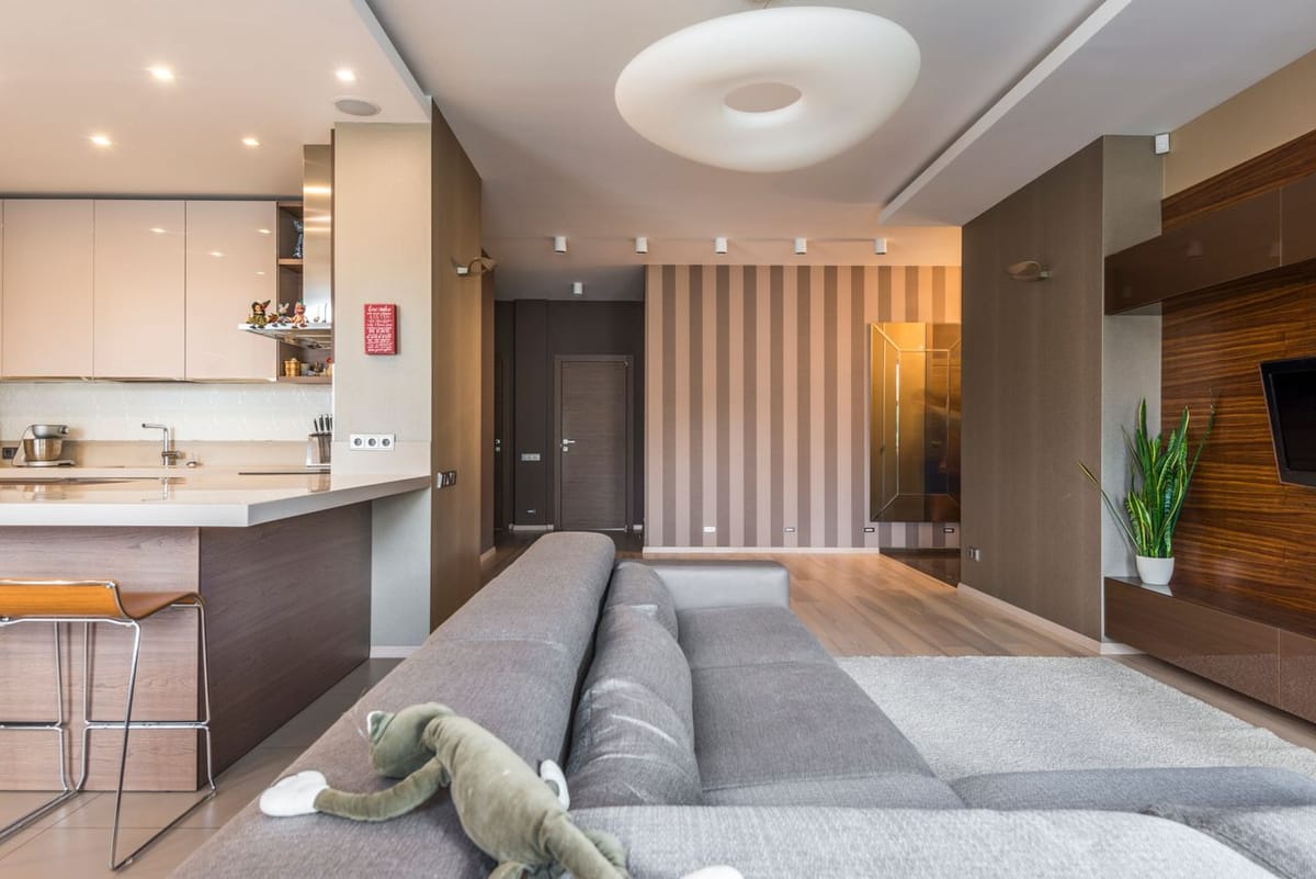An open-plan living room and kitchen with a grey sectional sofa, striped accent wall, kitchen island, and a wall-mounted TV.