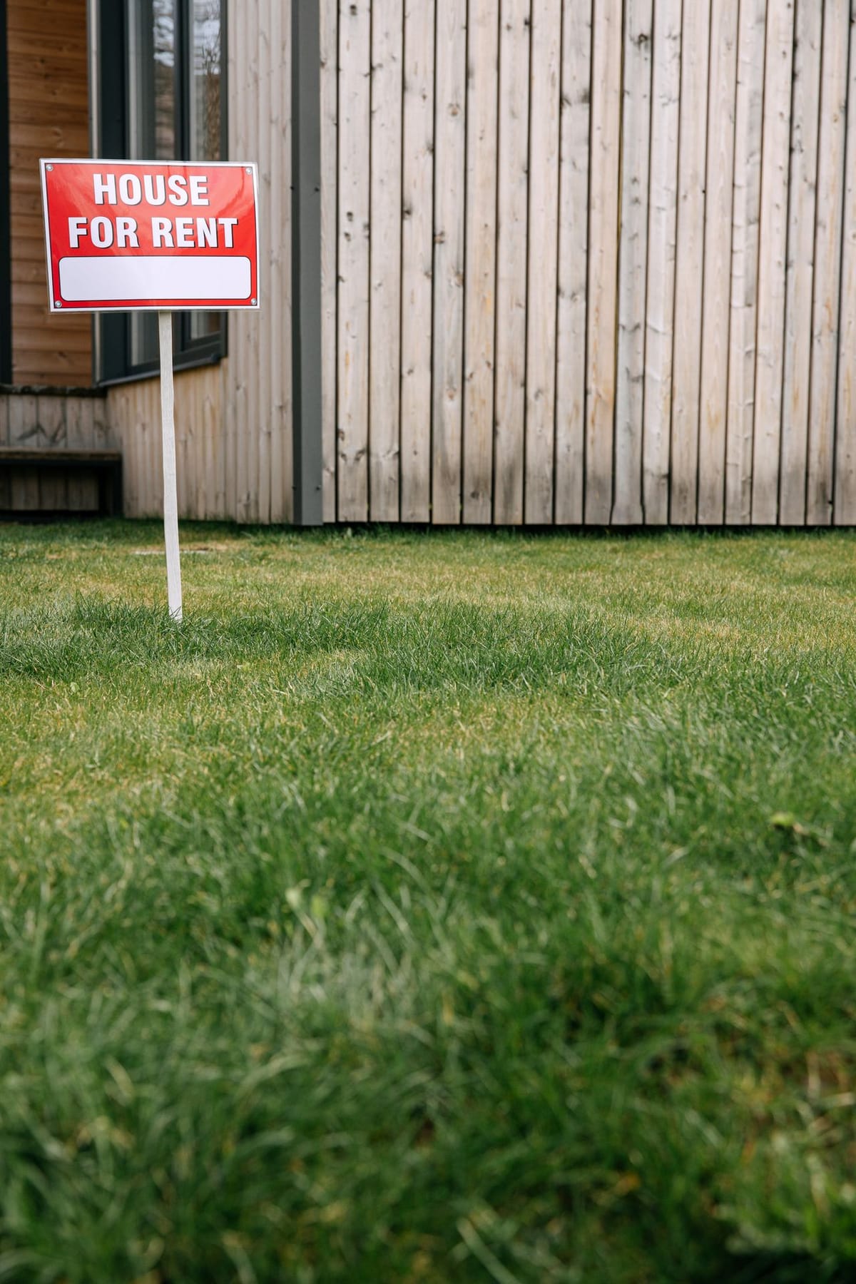 A red House For Rent sign on a white stake planted in a green lawn in front of a light wood-clad house exterior.
